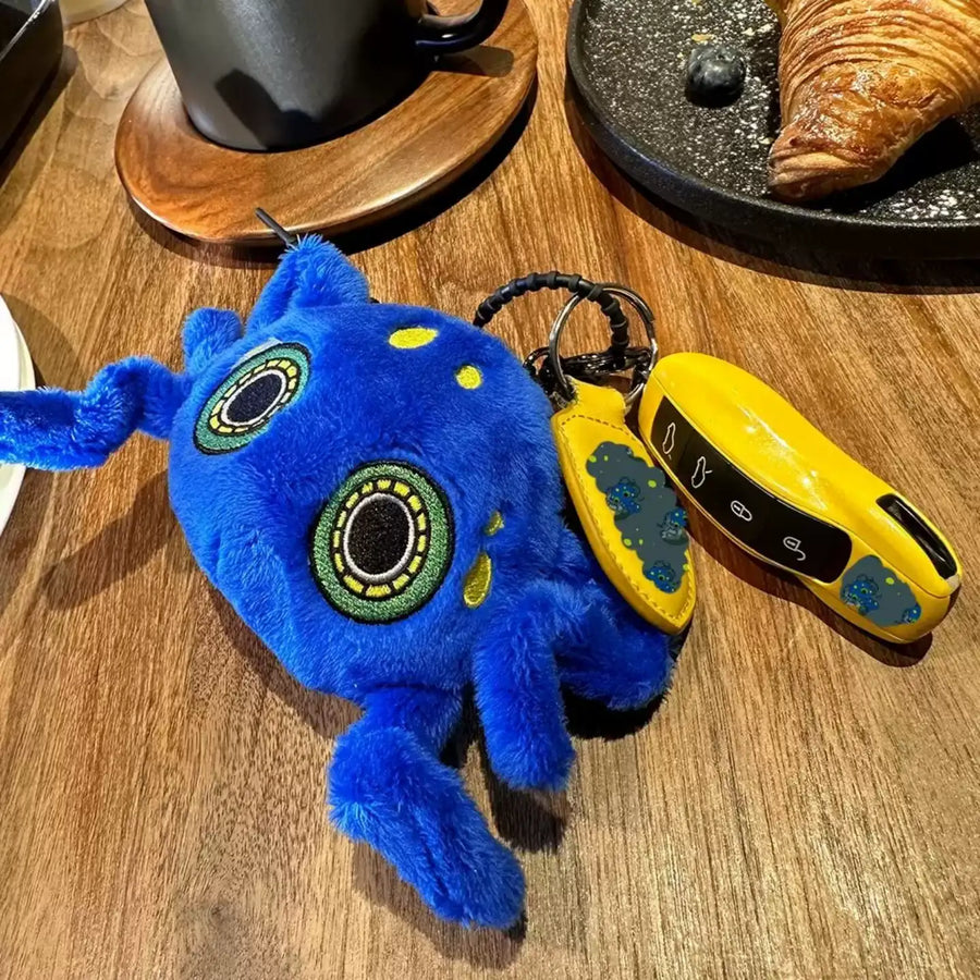 Blue Dark Envoy Octopus plush charm with green-and-yellow embroidered eyes and curling tentacles beside yellow keys on a cafe table