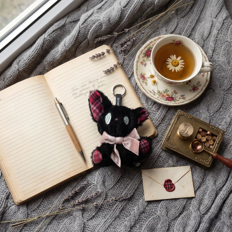 Eavesdropping Kitten black plush charm beside an open journal and floral teacup on a gray knitted blanket