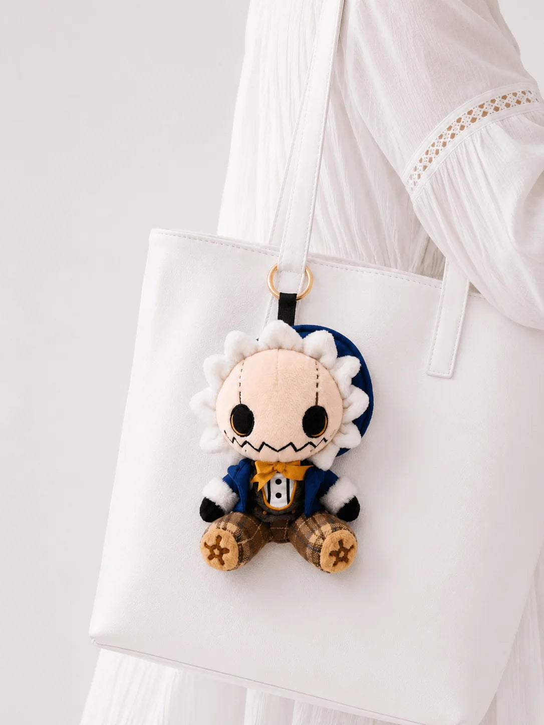 Elliott scarecrow plush charm hanging from a white tote, with a blue hat, mustard bow tie and plaid legs