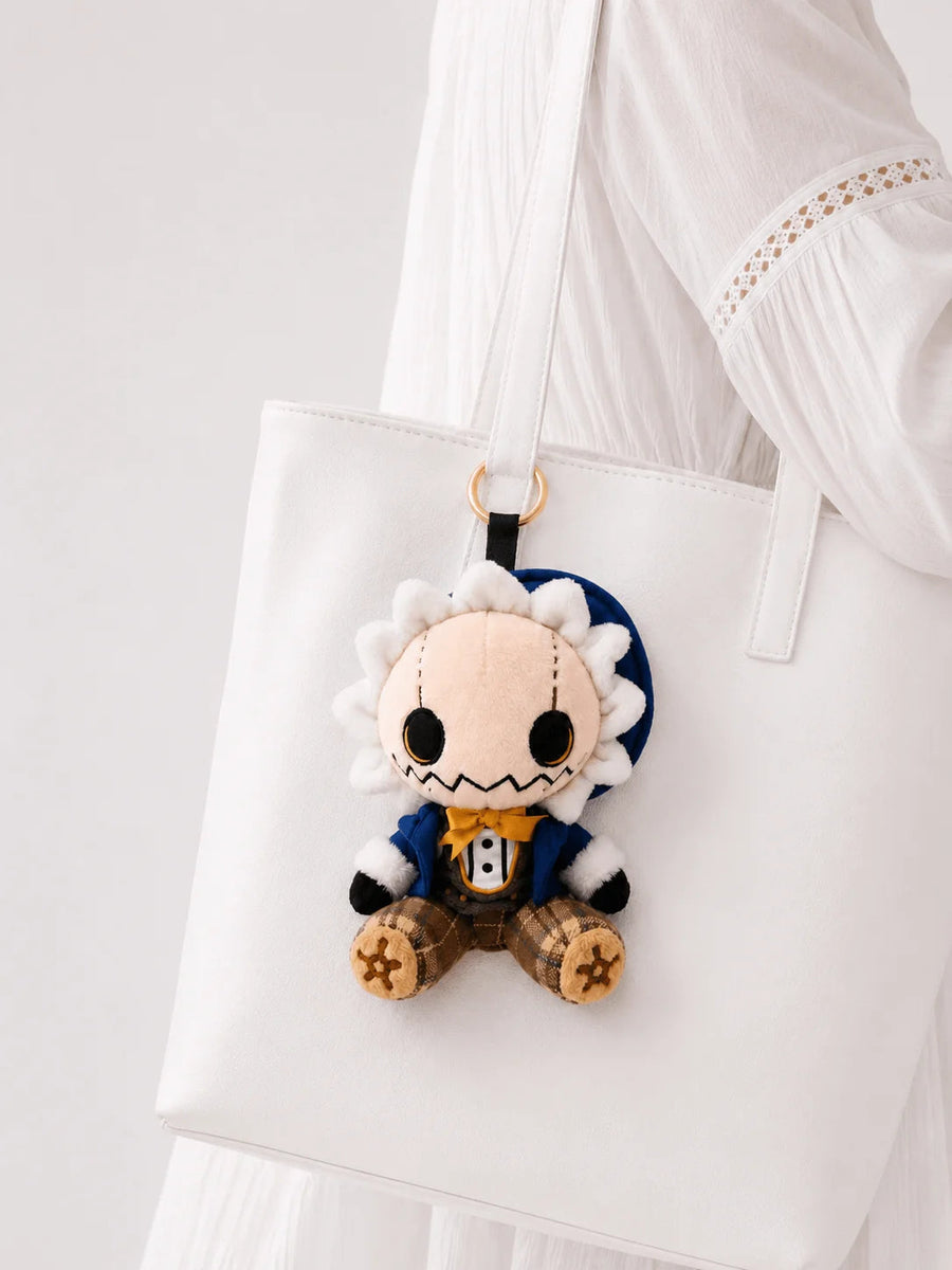 Elliott scarecrow plush charm hanging from a white tote, with a blue hat, mustard bow tie and plaid legs