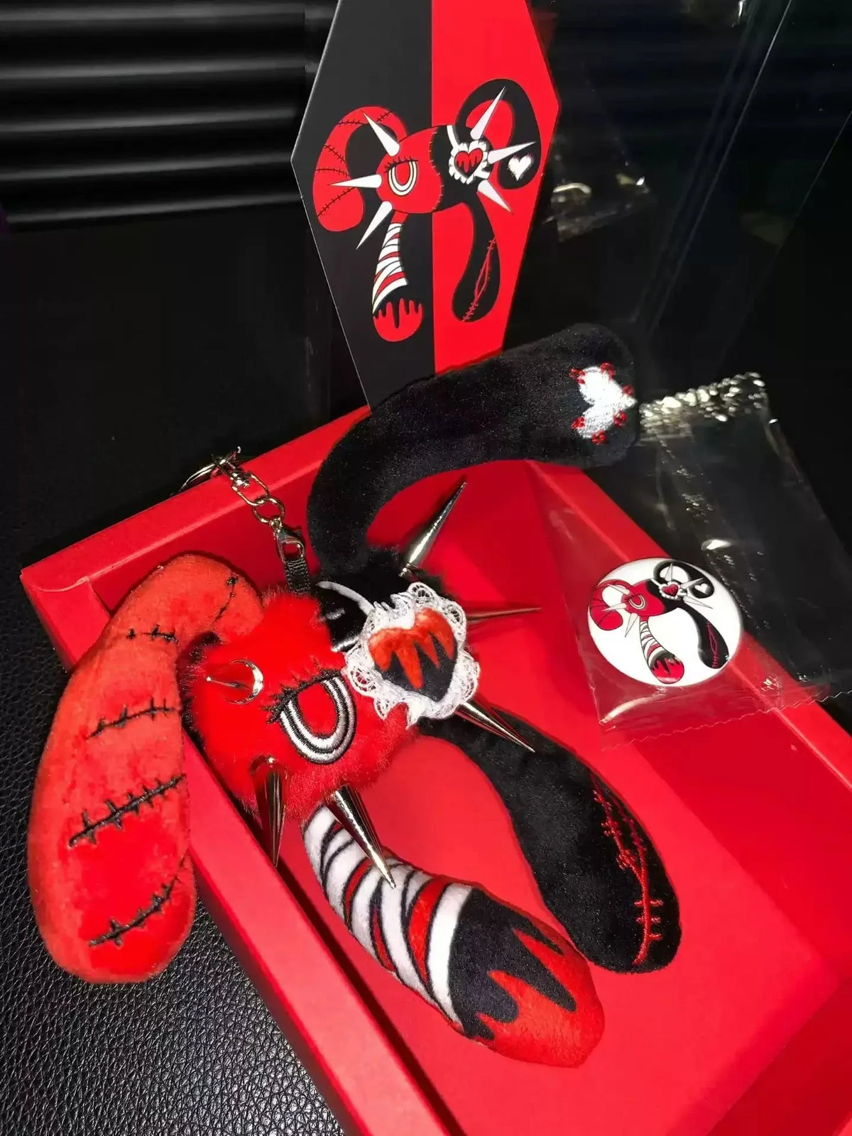 Rivet Rabbit red-and-black plush charm with its eye patch, displayed in a red gift box beside a badge and illustrated card