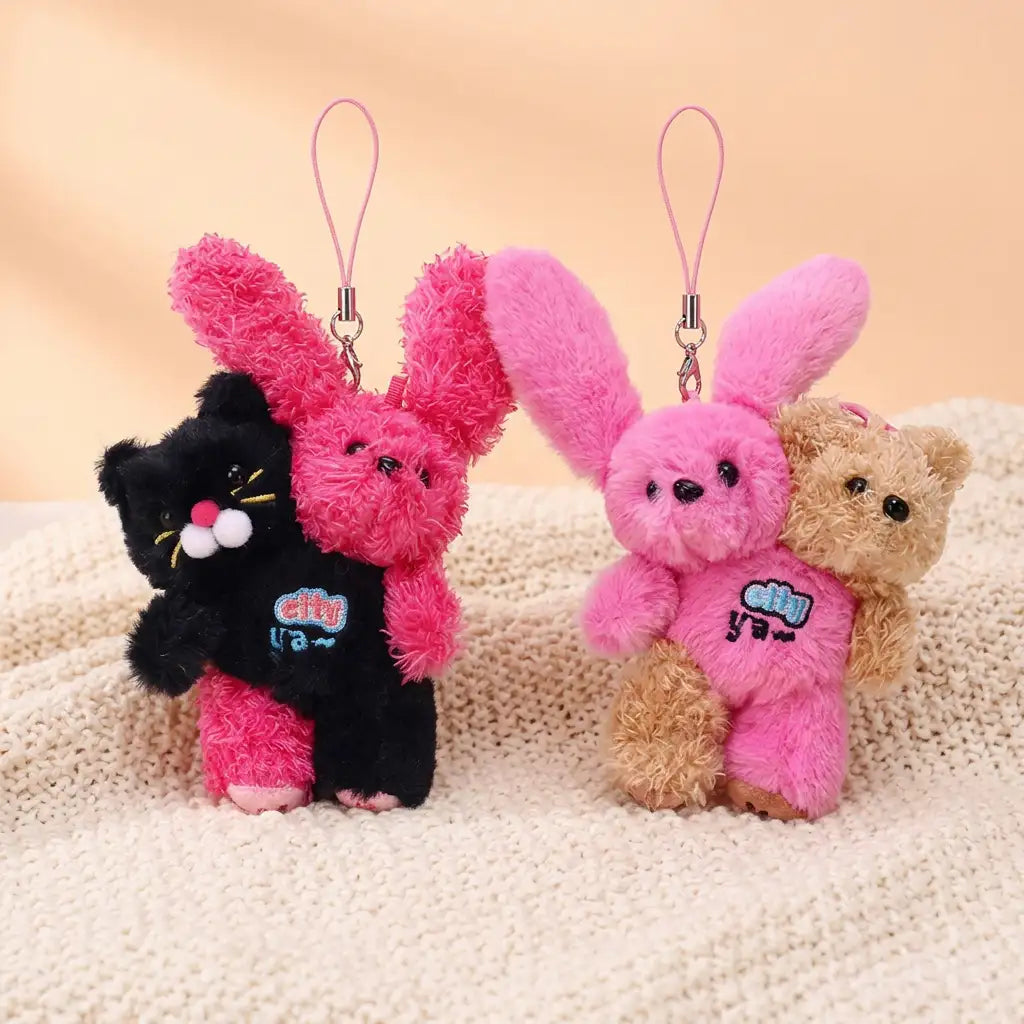 Two hugging plush charms standing side by side on a knit blanket: a black cat holding a curly hot-pink bunny, and a pink bunny held from behind by a honey-tan teddy bear, each with a hanging strap loop.