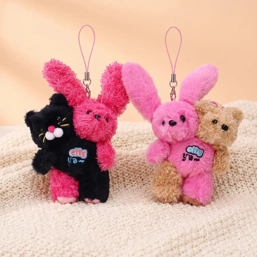 Two hugging plush charms standing side by side on a knit blanket: a black cat holding a curly hot-pink bunny, and a pink bunny held from behind by a honey-tan teddy bear, each with a hanging strap loop.