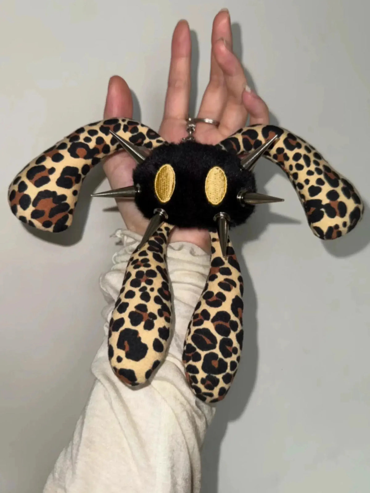 A hand holding a plush keychain shaped like a black spider with leopard-print legs and gold oval eyes, set against a plain light gray background.
