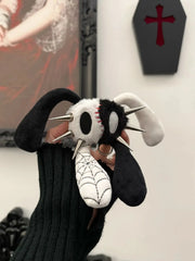A person wearing a black ribbed sweater holds a black-and-white plush bunny toy with metal spikes and a white spiderweb-patterned nose in a gothic-themed room.