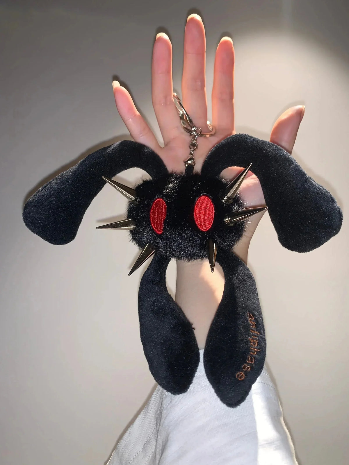 A black plush keychain shaped like a spiky monster with red eyes, held up by a hand against a plain light gray background.