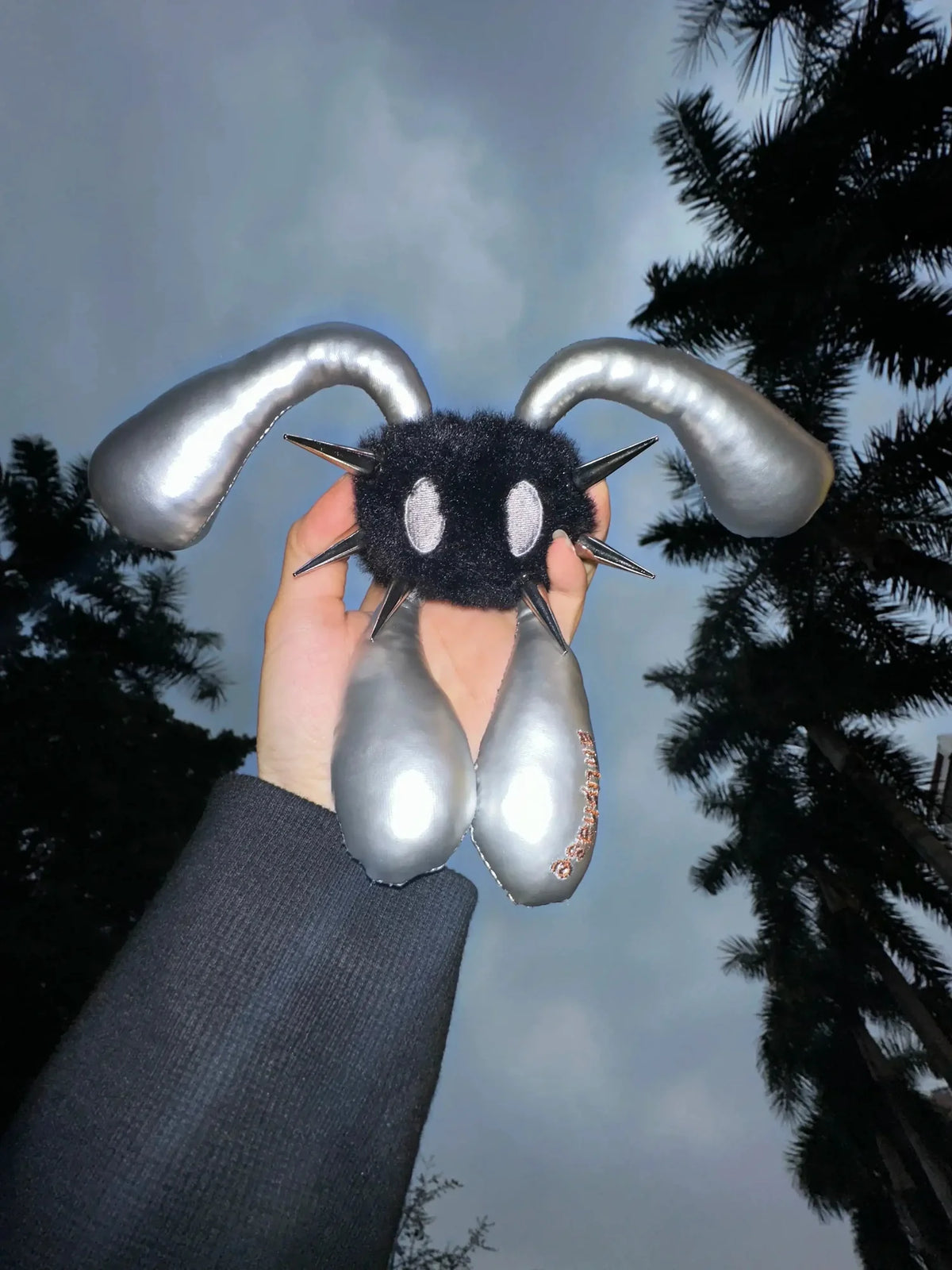 A hand holds a black fuzzy monster plush toy with long curved silver ears and metallic silver dangling elements against an outdoor night sky with trees.