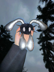 A hand holds a black fuzzy monster plush toy with long curved silver ears and metallic silver dangling elements against an outdoor night sky with trees.