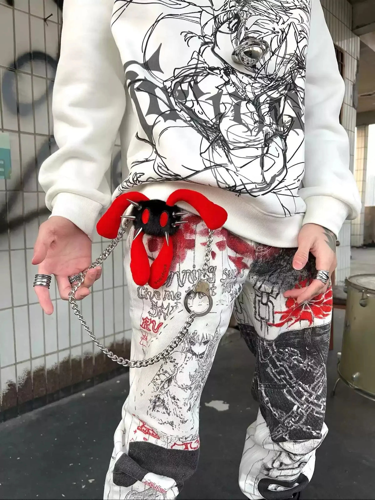 A person wearing a white graphic sweatshirt and matching white graphic pants with red and black graffiti-style print, holding a red plush toy with a silver chain in a real urban setting.