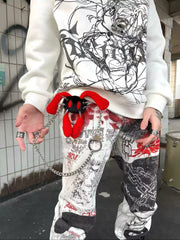 A person wearing a white graphic sweatshirt and matching white graphic pants with red and black graffiti-style print, holding a red plush toy with a silver chain in a real urban setting.