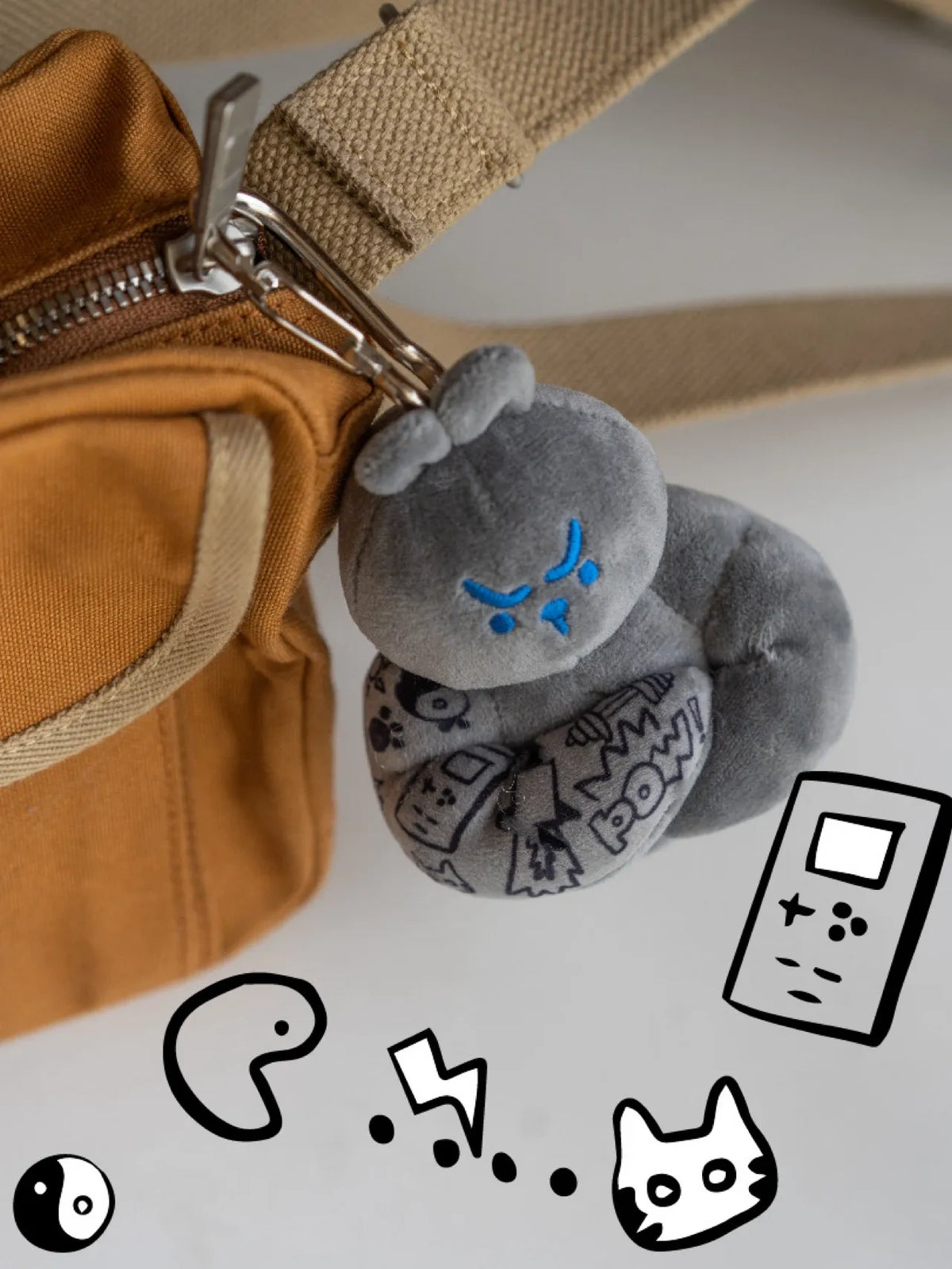 Grey muscle bunny plush bag charm clipped on tan crossbody bag strap with cartoon doodles, unisex fitness accessory