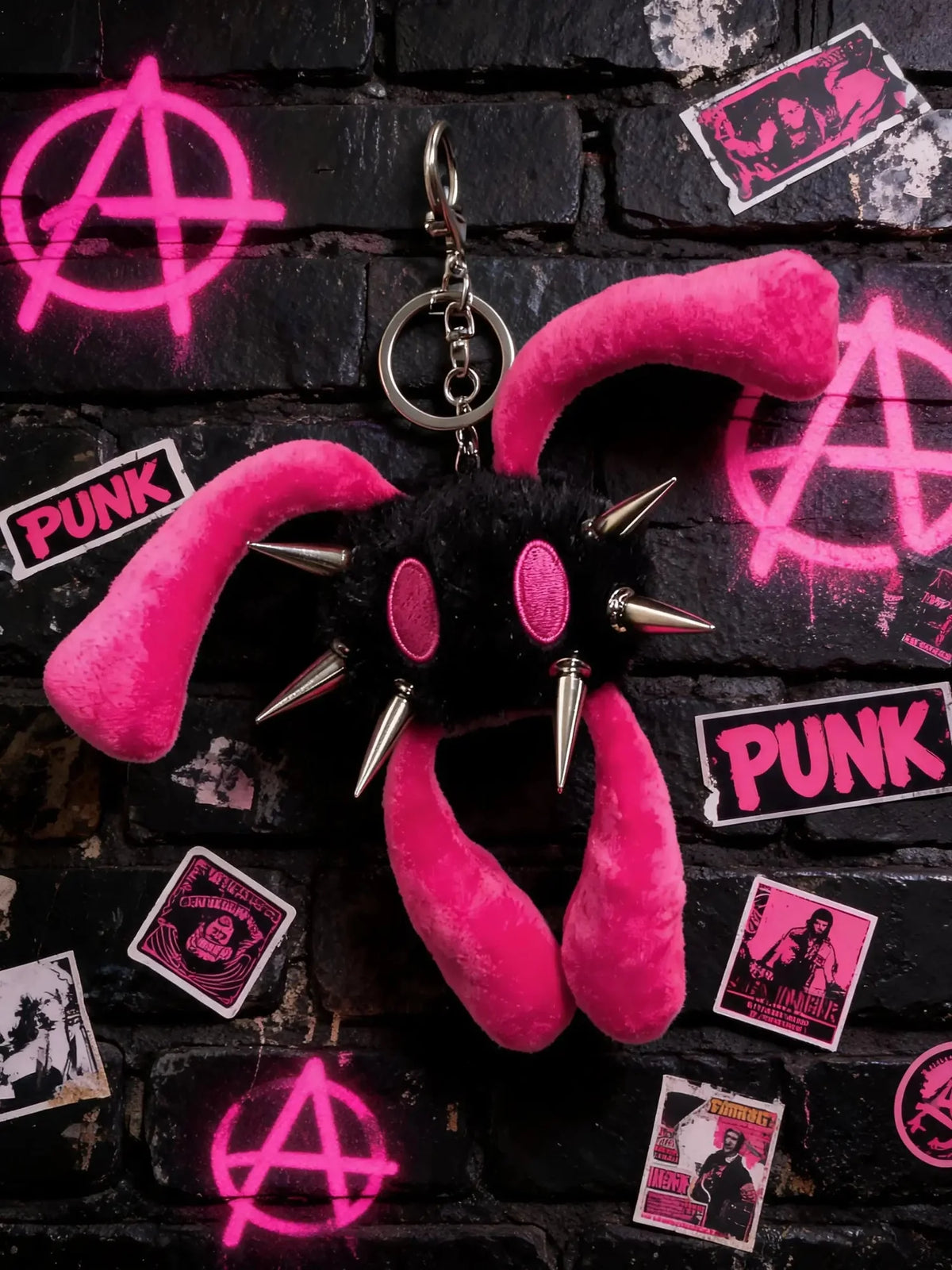 A pink and black punk-themed plush keychain with long floppy ears, metal spikes, and a silver key ring, displayed against a black brick wall with neon pink graffiti and sticker decorations.