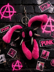 A pink and black punk-themed plush keychain with long floppy ears, metal spikes, and a silver key ring, displayed against a black brick wall with neon pink graffiti and sticker decorations.
