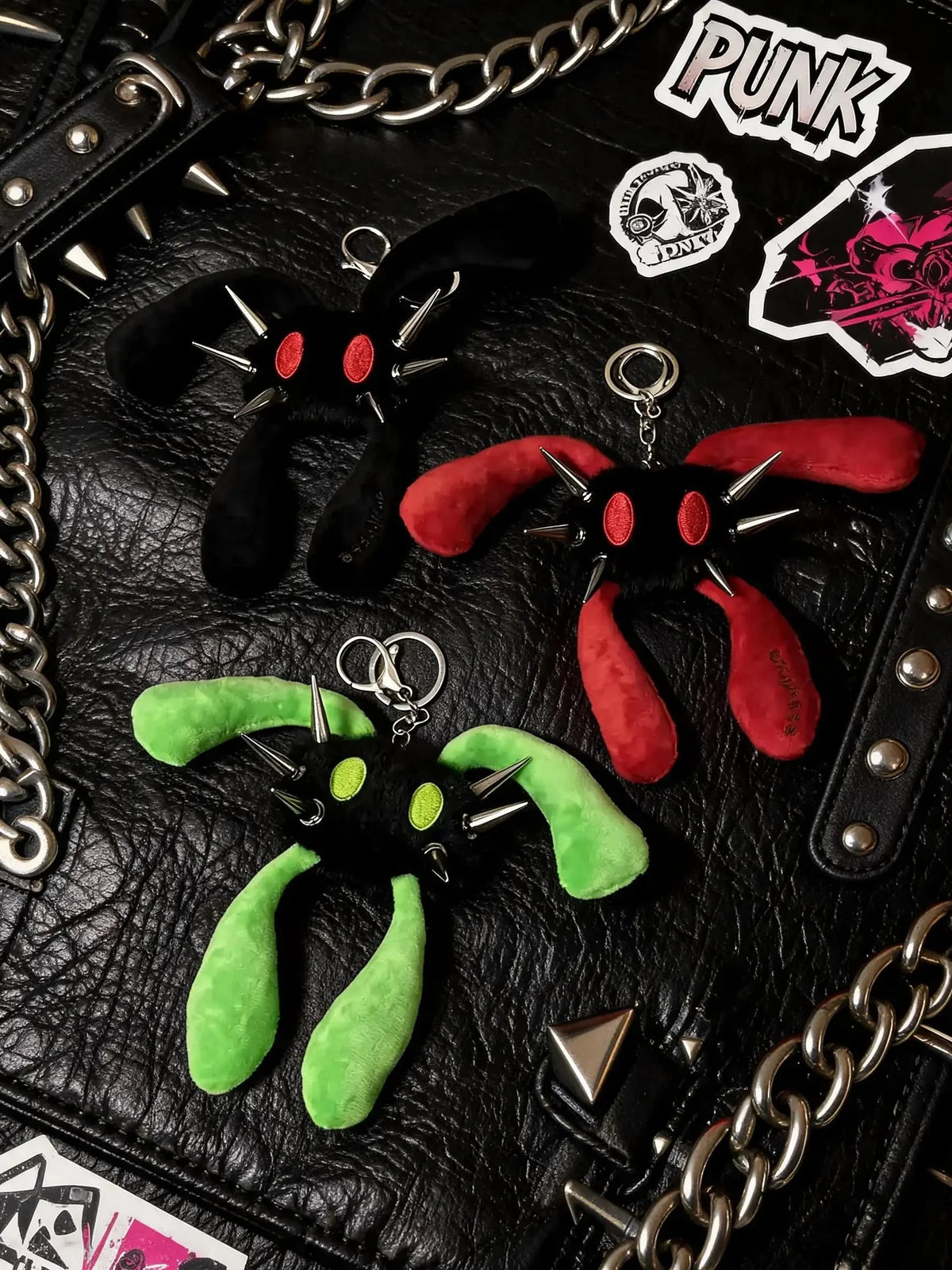 Three plush keychains shaped like spiky-eyed creatures in black, red, and green, arranged on a black leather surface with chains and stickers.