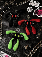 Three plush keychains shaped like spiky-eyed creatures in black, red, and green, arranged on a black leather surface with chains and stickers.