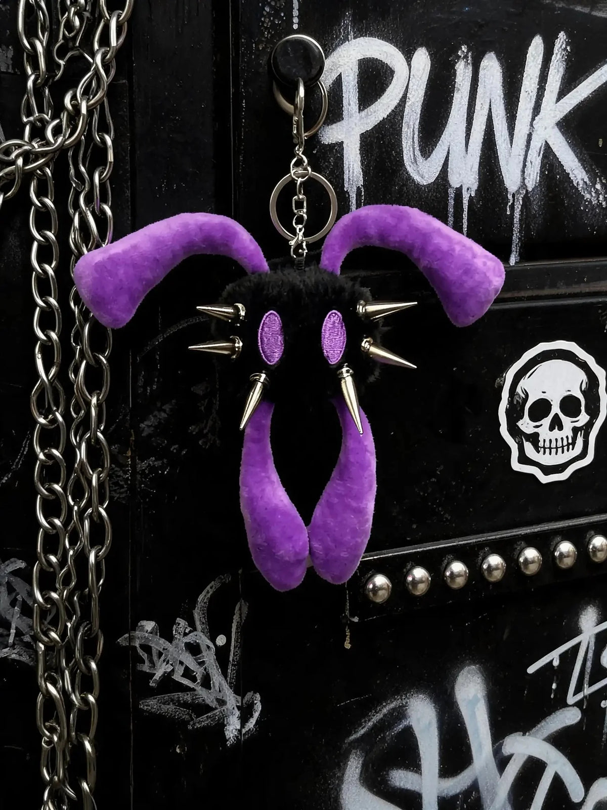 Purple and black plush monster keychain with long ears, metal spikes, and purple oval eyes, hanging on a black graffiti-covered surface with silver chains.