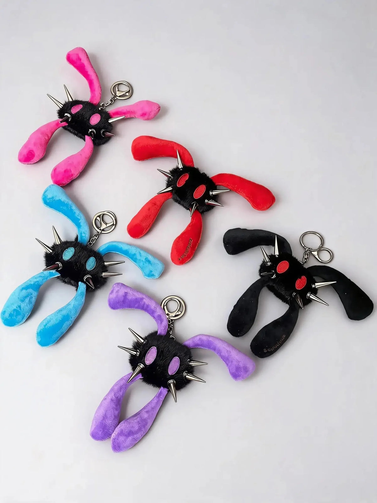 Five plush monster keychains with long fuzzy bunny-like legs and metal spikes, shown in pink, red, blue, purple, and black on a light gray background.