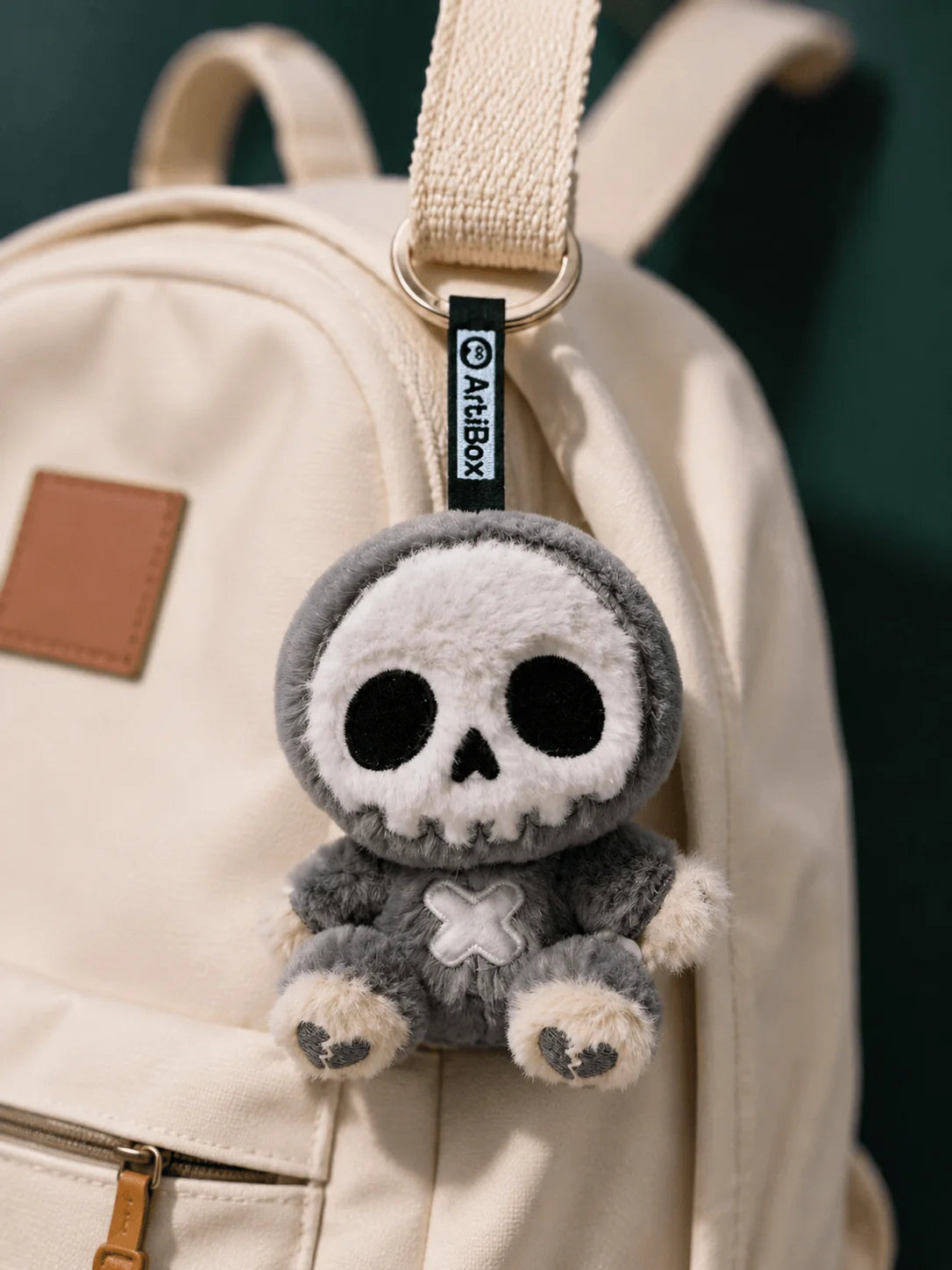 Little Angu plush charm hanging from a beige backpack, featuring a cute gray skull design.