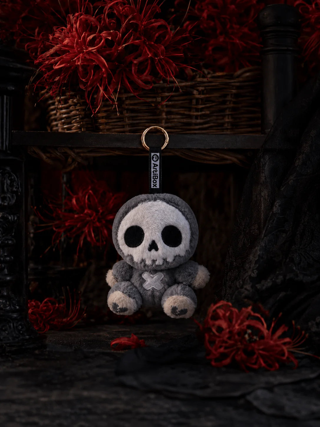 Little Angu plush charm in smoky-gray with oversized skull face and crossbone detail, surrounded by red decorations.