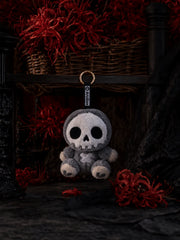 Little Angu plush charm in smoky-gray with oversized skull face and crossbone detail, surrounded by red decorations.